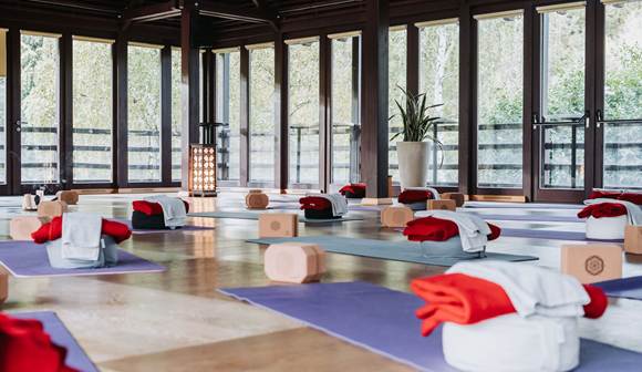 Körper in Bewegung & Geist im Gleichgewicht Yogaraum mit Glasfront, auf dem Boden diverse Yogamatten, Hocker und Handtücher.