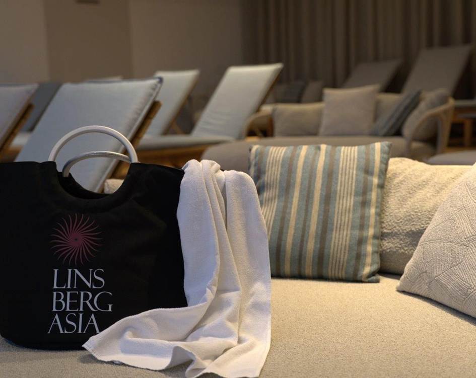 Badetasche mit Bademantel im Premium Day Spa Linsberg Asia Schwarze Badetasche mit Linsberg Asia Logo und weißem Bademantel liegt auf einer Liege im Ruheraum mit weiteren Liegen im Hintergrund.