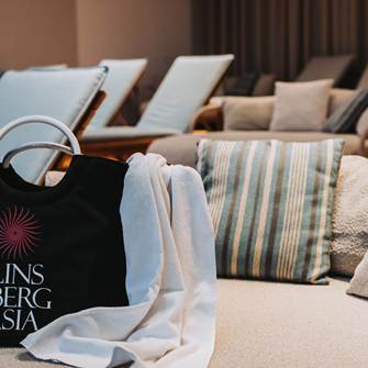 Genießen Sie einen Wellnesstag im Linsberg Asia Badetasche der Therme Linsberg Asia aus er ein Handtuch rausschaut auf einer Liege.